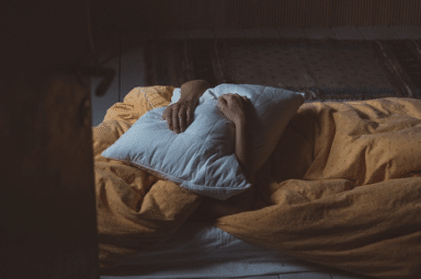 Person lying in bed under a yellow duvet, holding a white pillow over their head in a dimly lit room.